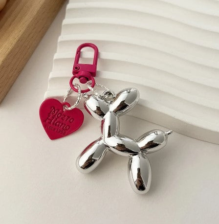 A metallic balloon dog keychain with a pink heart charm and clasp, displayed on a textured surface.