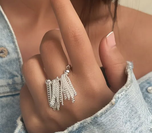 A silver tassel ring with cascading chain details, worn on a hand styled with denim for a trendy and edgy look.