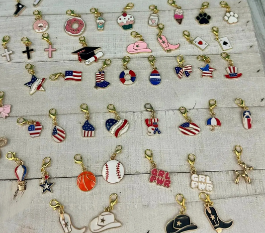 A collection of assorted clip-on shoe charms with gold-tone clasps, displayed on a white wooden background, featuring patriotic, western, sports, and trendy designs.

