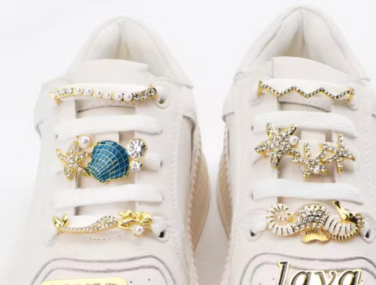 A pair of white sneakers decorated with gold-tone lace-through shoe charms featuring rhinestones, pearls, and ocean-inspired designs.

