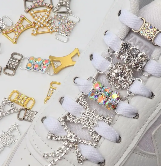 A pair of white sneakers decorated with lace-through rhinestone shoe charms, featuring crystal-encrusted designs and metallic accents.

