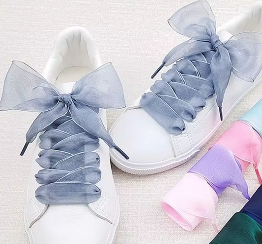 A pair of white sneakers laced with sheer ribbon shoelaces tied into bows, with additional pastel-colored ribbon laces displayed beside them.

